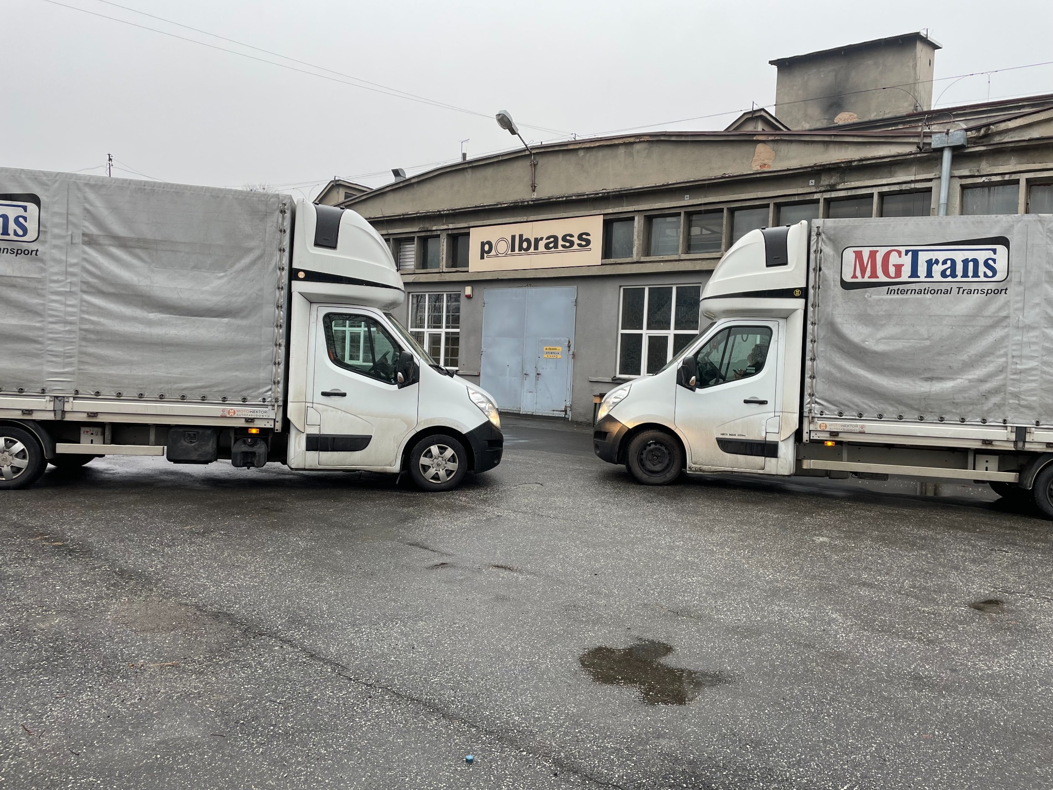 Dwa białe busy dostawcze, jeden z logo MGTrans, stojące naprzeciwko siebie na mokrym asfalcie w Sosnowcu, z budynkiem firmy Polbrass w tle.