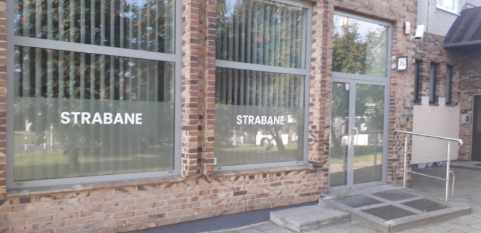Fasada budynku z cegły z dużymi oknami i szarymi ramami, na oknach naklejone napisy 'STRABANE', szklane drzwi wejściowe z rampą dla niepełnosprawnych.