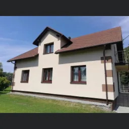 Otynkowany i pomalowany dom jednorodzinny z brązowymi oknami i dachem z gontu bitumicznego, widoczny fragment balkonu z metalową barierką.