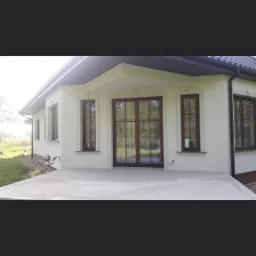 Wykończona elewacja domu jednorodzinnego z brązowymi oknami PCV z podziałami szprosowymi, jasna elewacja, widoczne orynnowanie.