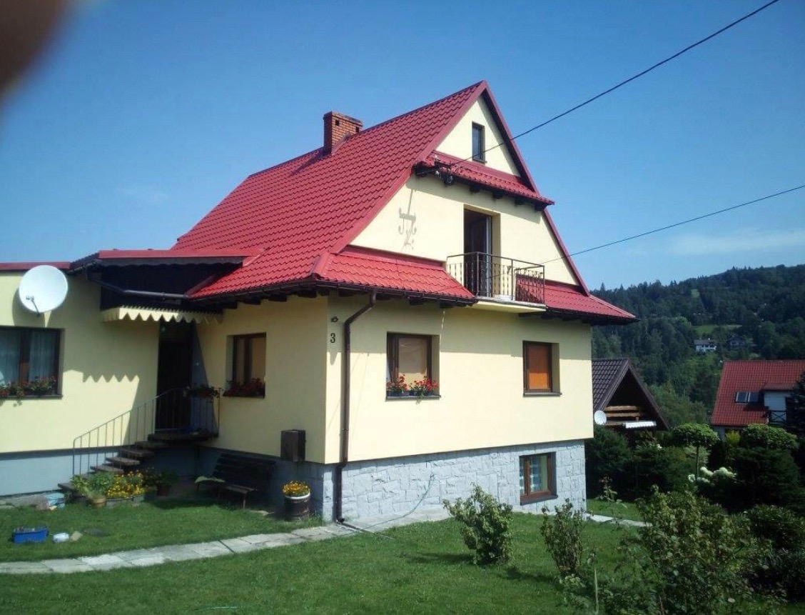 Żółty dom jednorodzinny z czerwoną dachówką, oknami z kwiatami i wykończonym kamieniem fundamentem, na tle zielonego ogrodu i zalesionych wzgórz.