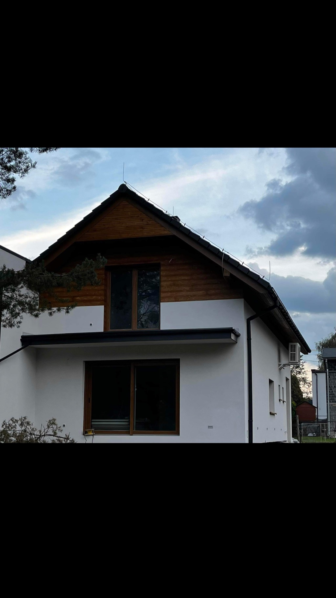 Nowoczesny dom jednorodzinny z białymi ścianami i drewnianym wykończeniem pod dachem, widoczne brązowe okna i klimatyzator na bocznej ścianie, pochmurne niebo w tle.