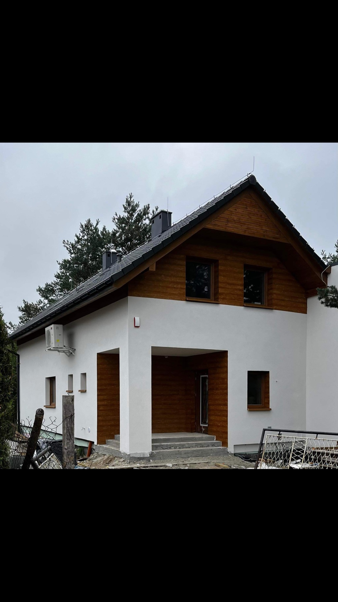 Nowoczesny dom jednorodzinny z białą elewacją i drewnianym wykończeniem pod dachem, widoczne okna z brązowymi ramami, klimatyzator na ścianie i prowizoryczne ogrodzenie na pierwszym planie.