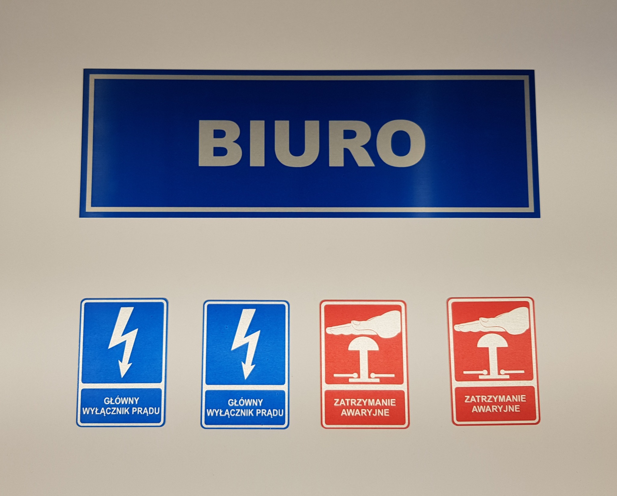 Oznakowanie biura: niebieski szyld z napisem 'BIURO' oraz naklejki informacyjne o głównym wyłączniku prądu i zatrzymaniu awaryjnym.