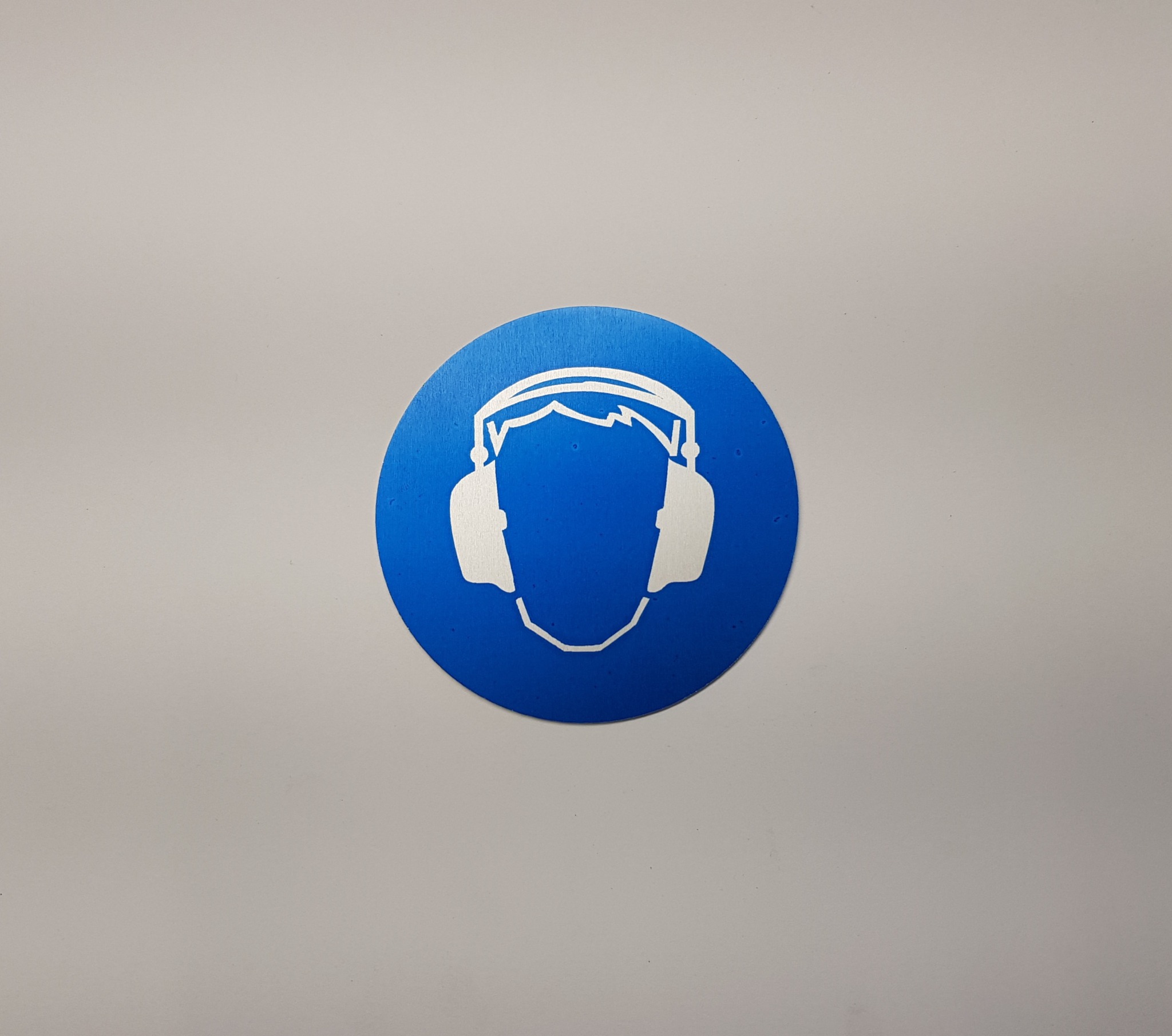 Niebieska okrągła naklejka z białym piktogramem osoby w słuchawkach ochronnych na szarym tle.