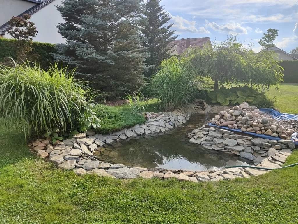 Oczko wodne z kamienną obudową i kaskadą, otoczone zielenią, w tle iglaste drzewo i budynek mieszkalny.