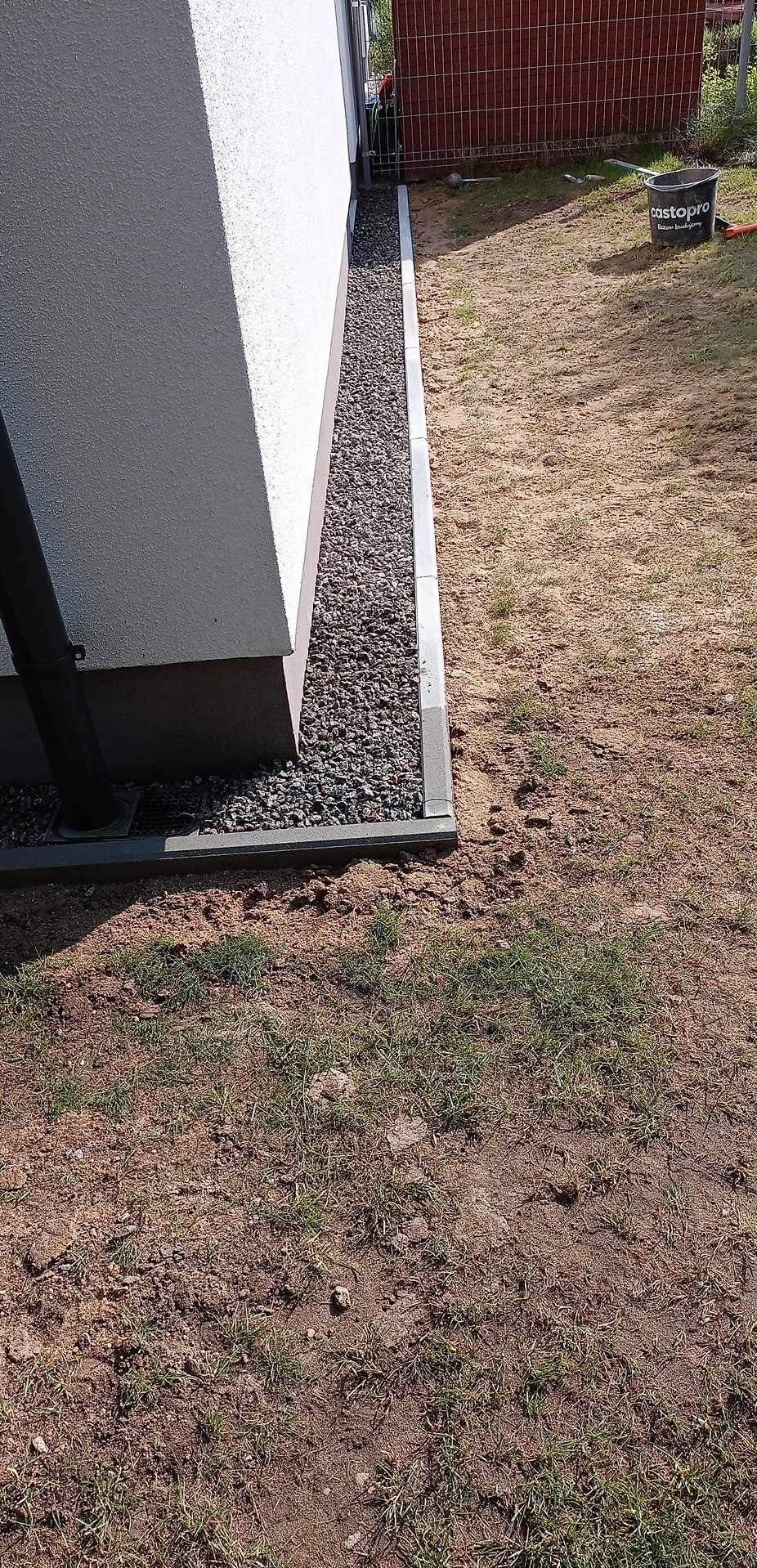 Wykończenie obrzeża domu z szarej kostki i wypełnienie przestrzeni szarym żwirem, widoczny fragment rynny spustowej, w tle wiadro budowlane.