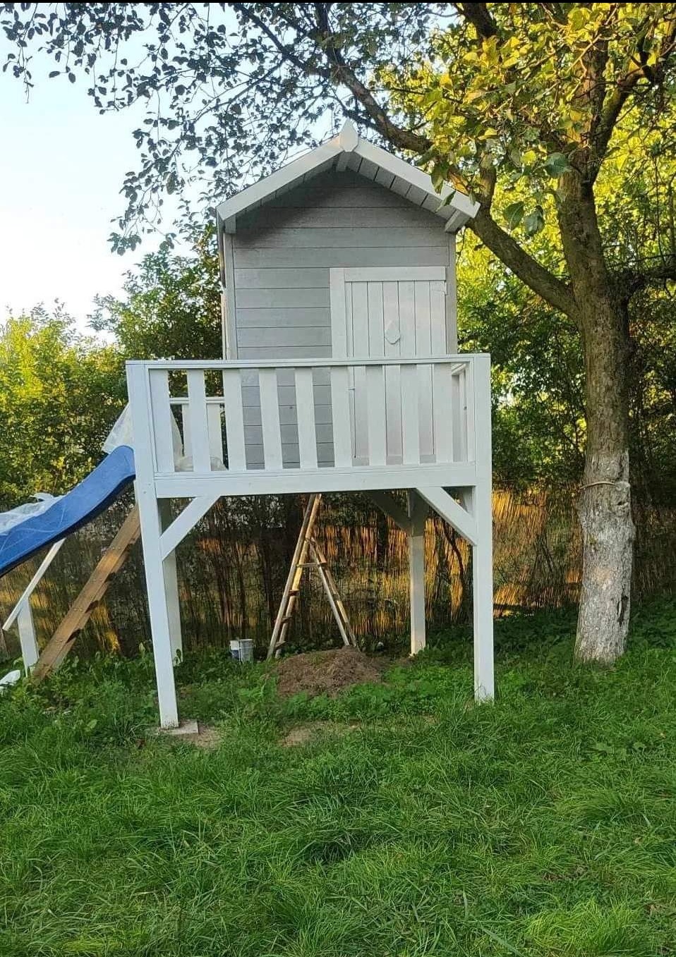 Domek na drzewie w ogrodzie, szaro-biała konstrukcja z drabinką i zjeżdżalnią, widok z poziomu trawy, w tle drzewa i krzewy.