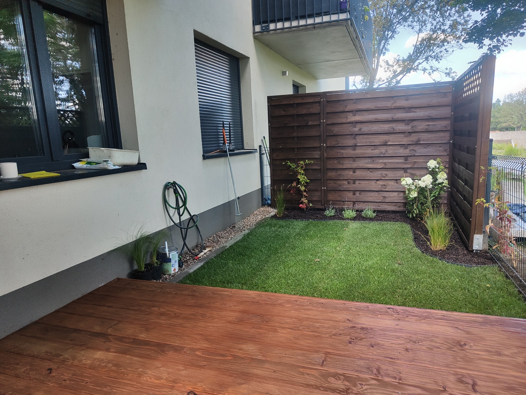 Niewielki, zagospodarowany ogródek przydomowy z drewnianym tarasem, świeżo posiana trawa, drewniane ogrodzenie i nasadzenia roślin ozdobnych.