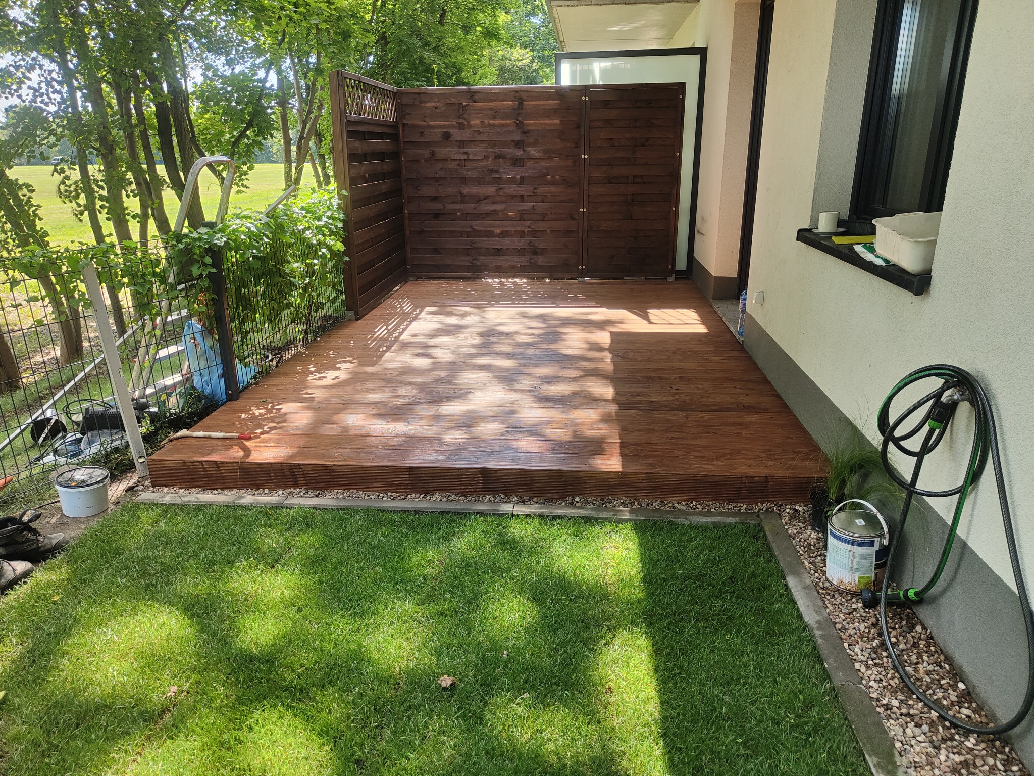Świeżo zaimpregnowany taras drewniany z ażurowym ogrodzeniem, widok na trawnik i drzewa w słoneczny dzień, obok ściany budynku z wężem ogrodowym i puszką farby.