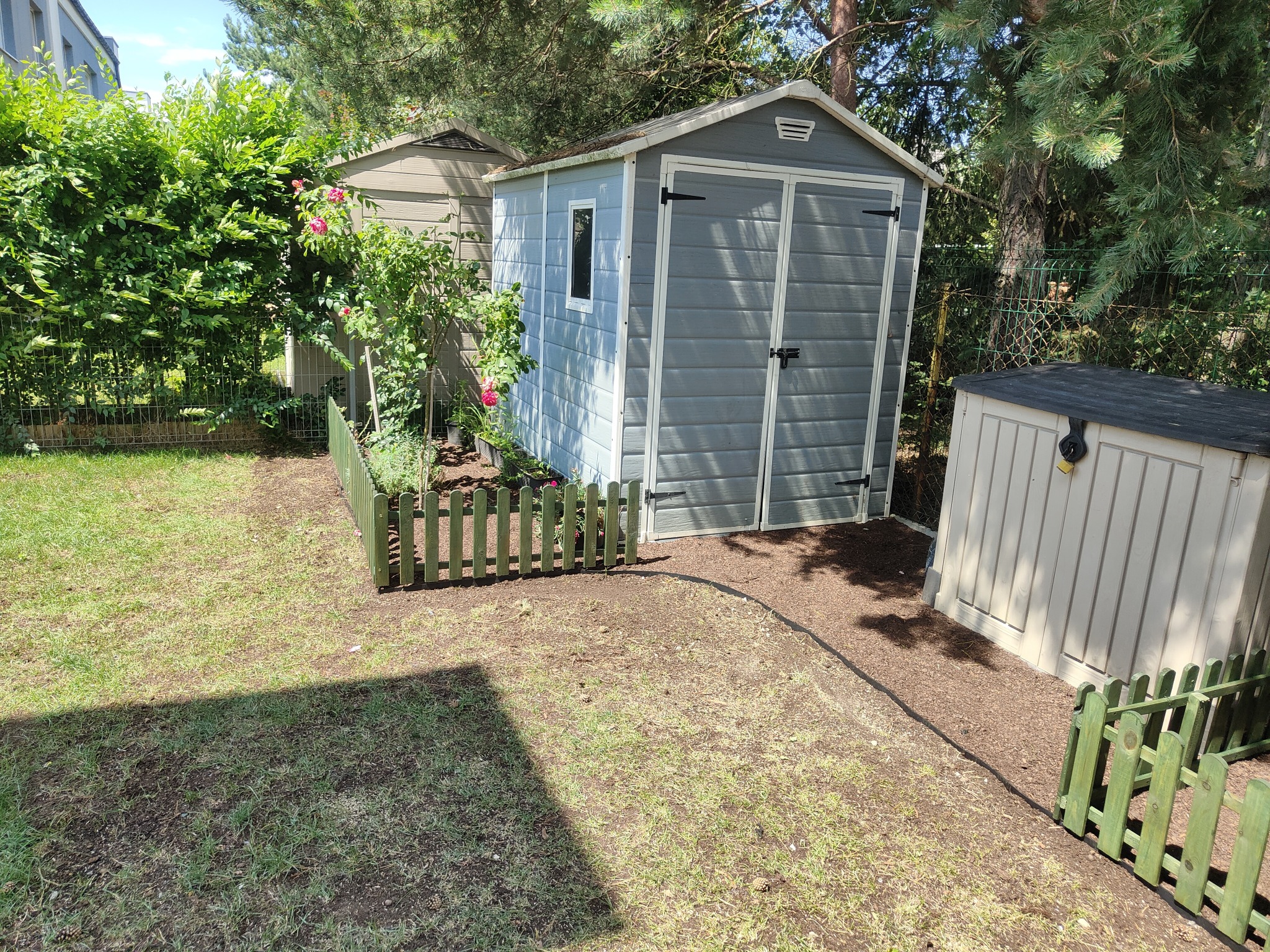 Dwa szare domki narzędziowe w ogrodzie, jeden z jasnoniebieskimi ścianami i czarnymi zawiasami, otoczony niskim, zielonym płotkiem, obok rabaty z kwitnącą różą.