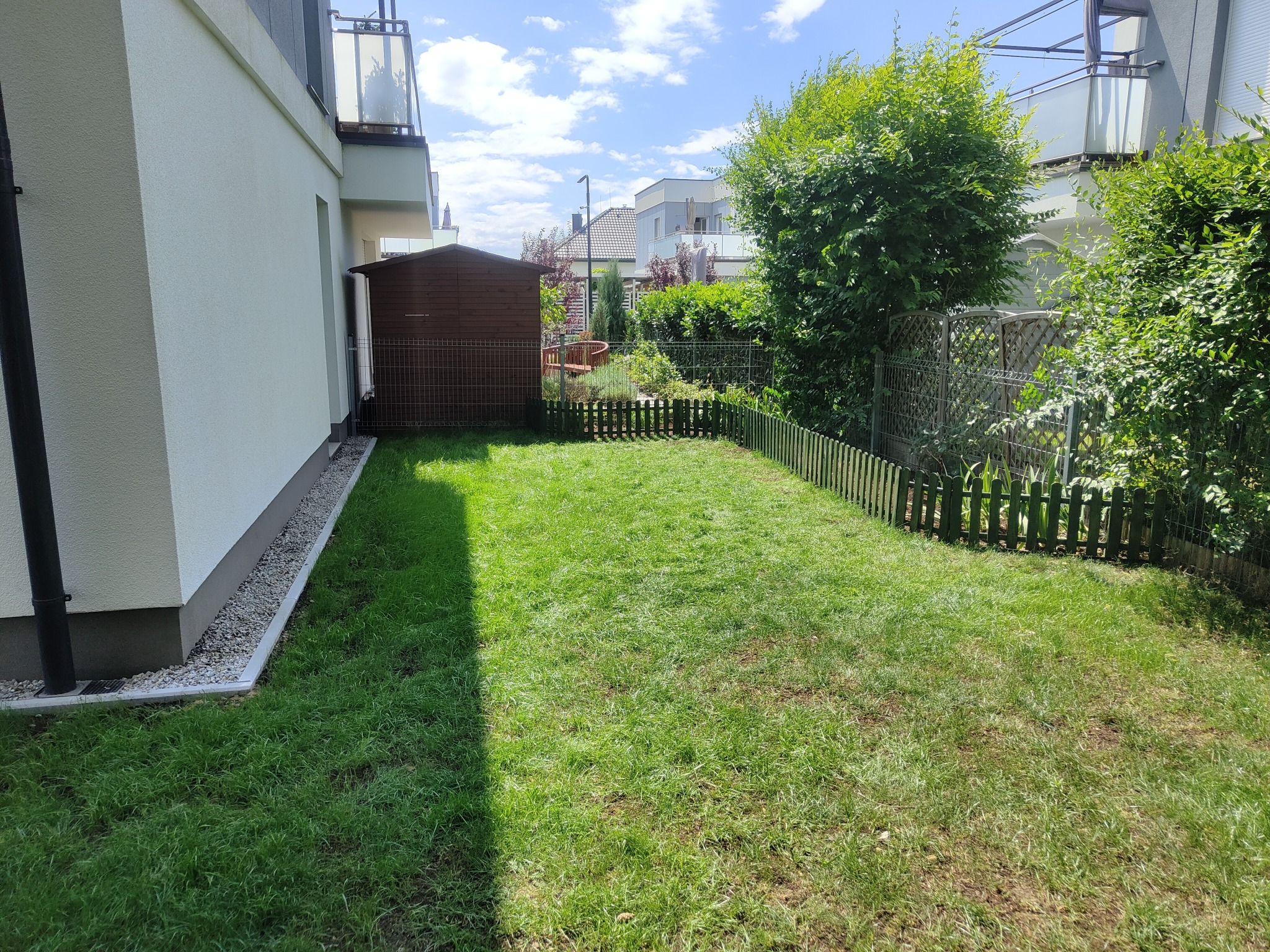 Widok na świeżo założony trawnik z rolki w przydomowym ogrodzie, z widoczną drewnianą szopą narzędziową i niskim płotkiem oddzielającym od sąsiedniej posesji.