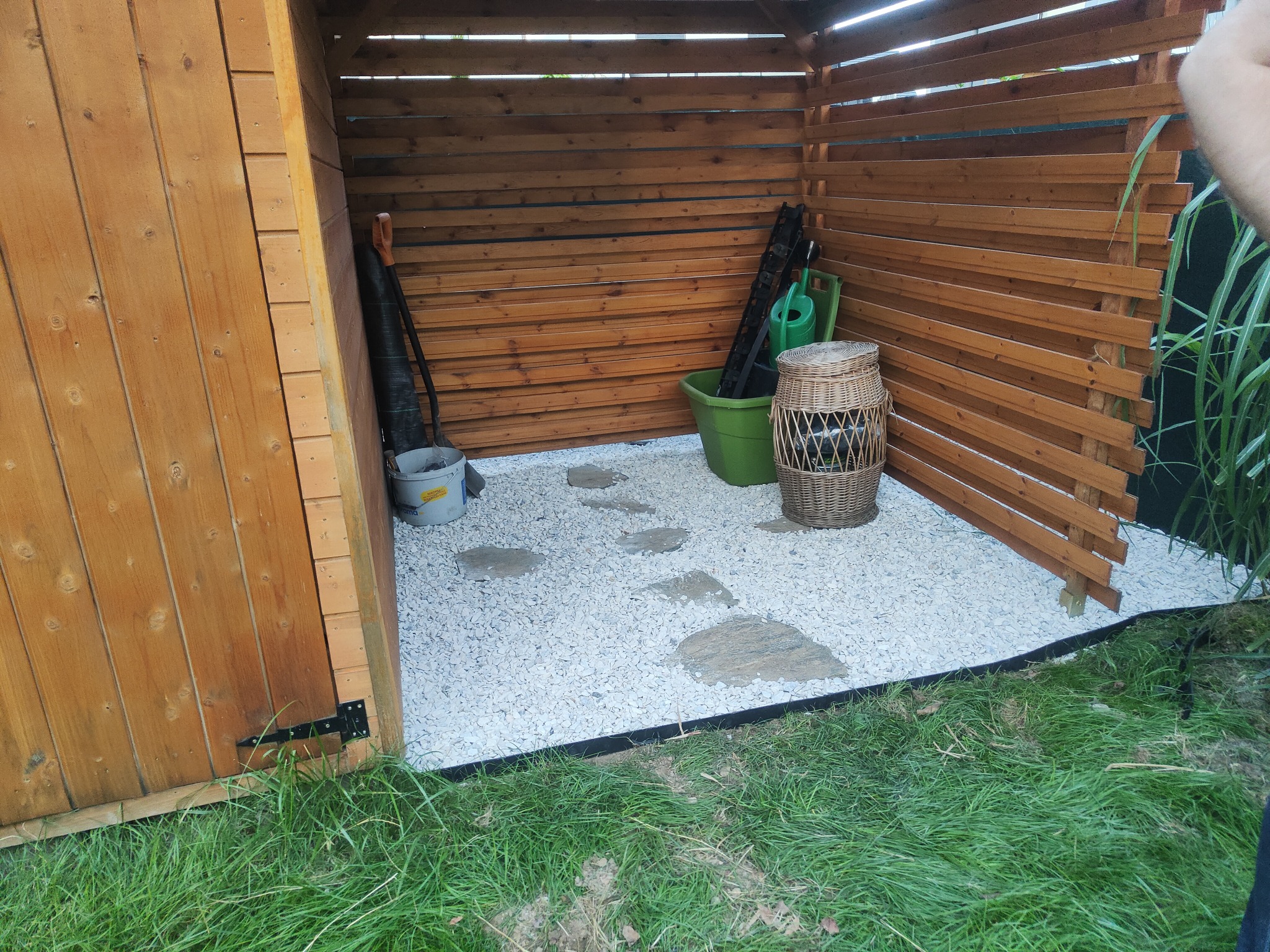 Zadaszona przestrzeń ogrodowa z drewnianymi ścianami, wysypana białym żwirem i kamiennymi płytami, z widocznymi narzędziami ogrodniczymi i dekoracyjnym koszem.