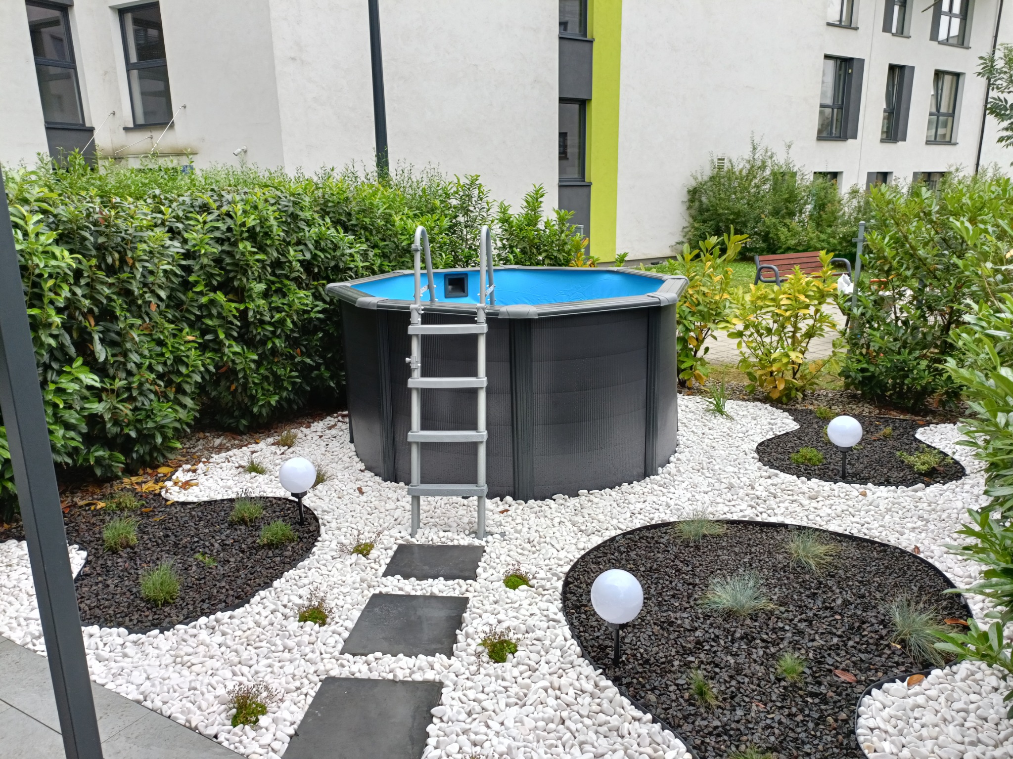 Ogród z basenem naziemnym otoczonym białymi kamieniami, czarnymi rabatami z roślinami i ścieżką z płyt betonowych.