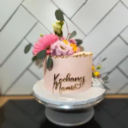 Różowy tort 'Kochanej Mamie' z kwiatami i złotymi płatkami, na paterze, tło z białych płytek. Dekoracja okolicznościowa.