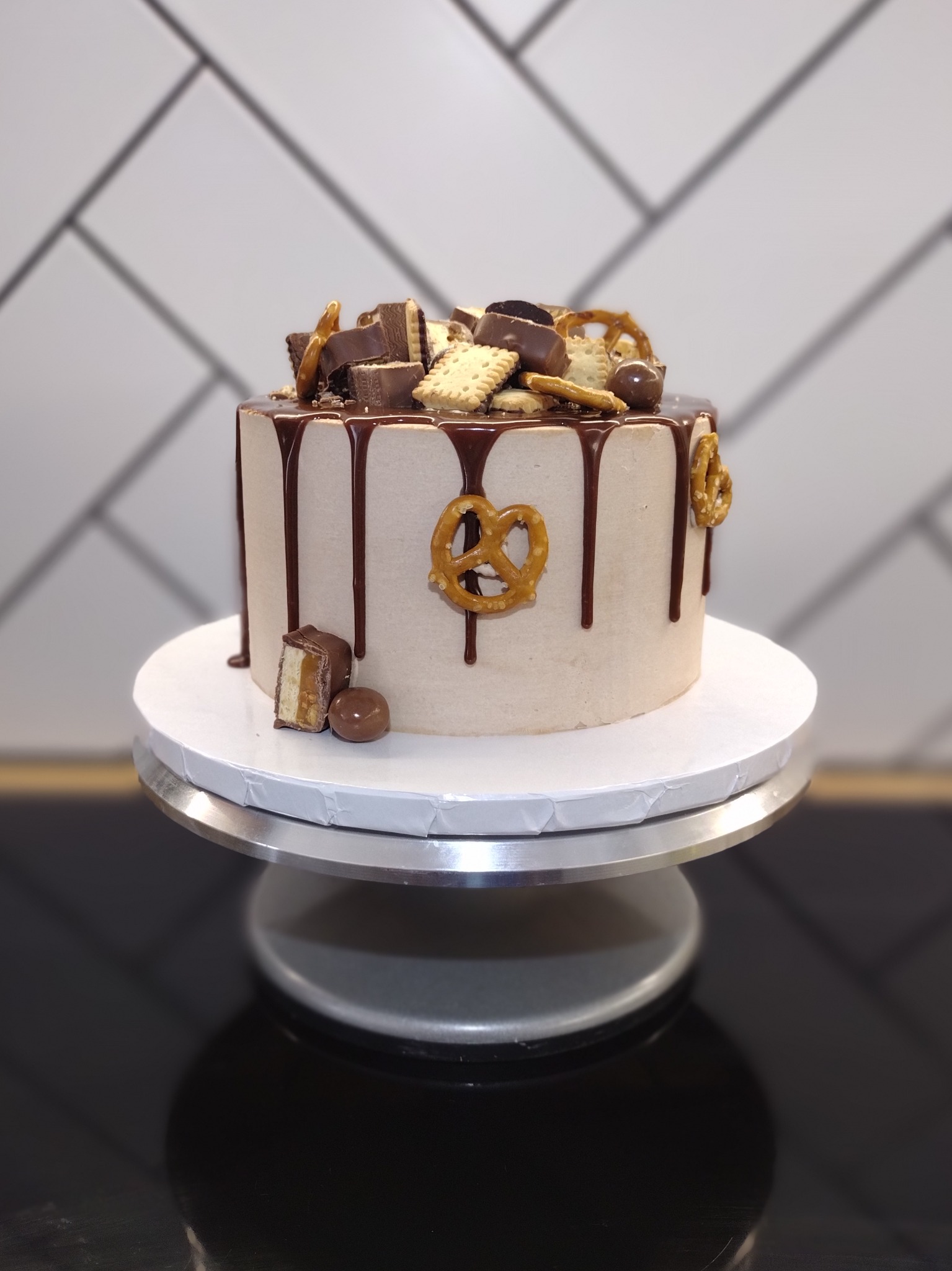 Tort z kremem, polewą czekoladową i dekoracjami z precelków, batoników i ciastek, ustawiony na srebrnej paterze na tle białych płytek.