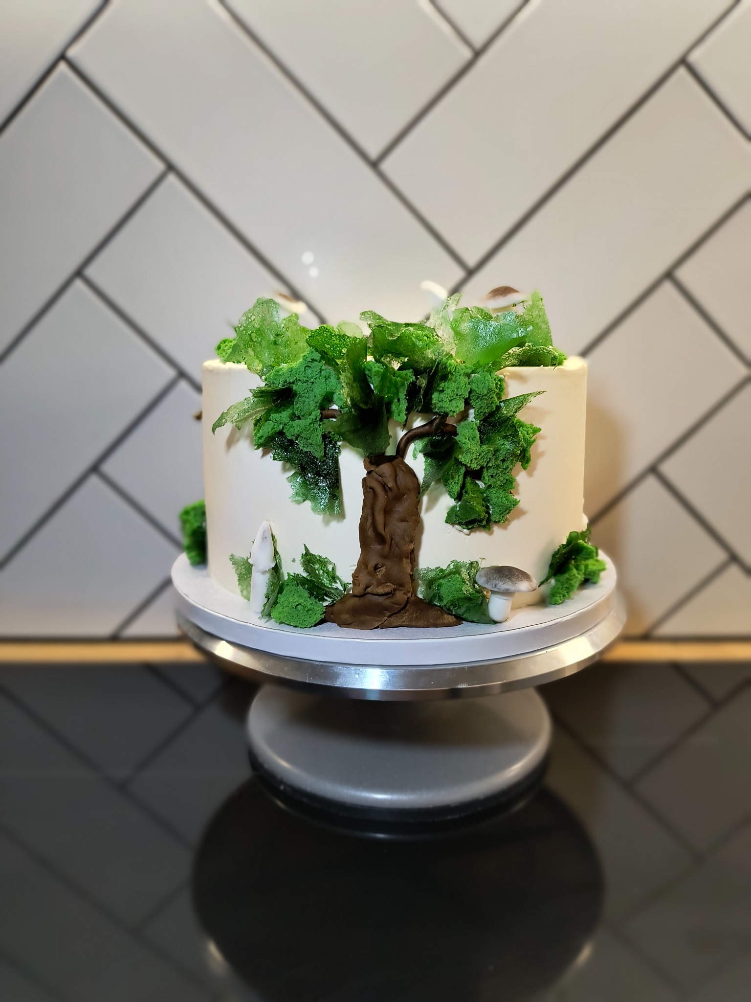 Tort urodzinowy z motywem leśnym: biały krem, brązowe drzewo z lukru, zielone liście z galaretki i marcepanowe grzyby na obrotowym stojaku.