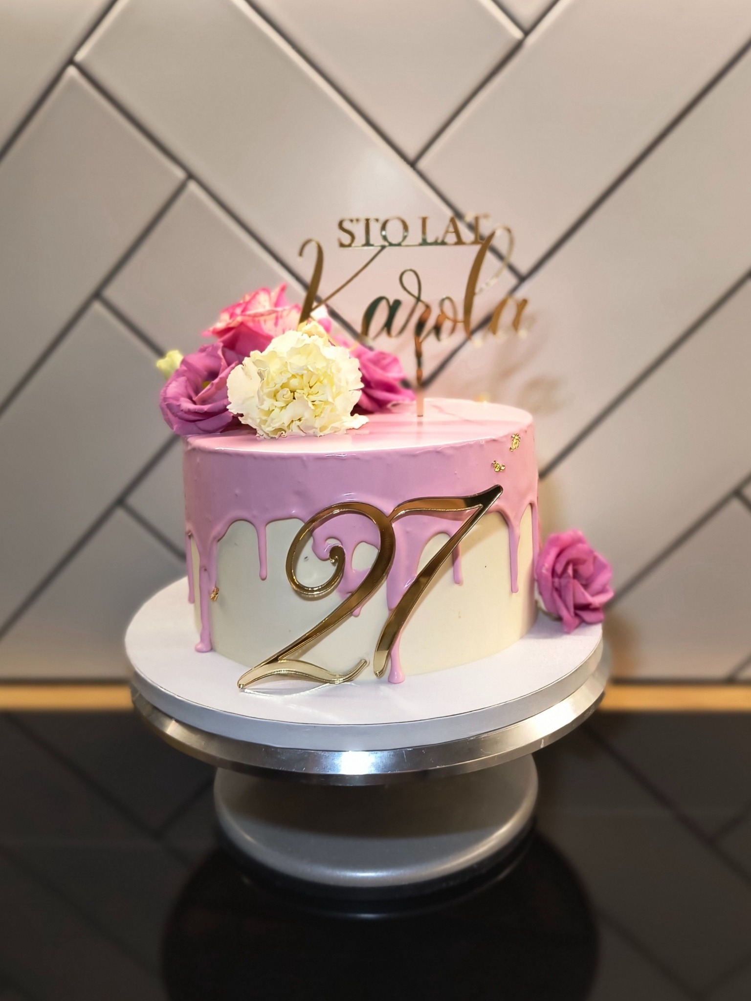 Tort urodzinowy z różowym lukrem, kwiatami i złotym napisem 'Sto lat Karola' oraz cyfrą 27, umieszczony na obrotowej podstawie.