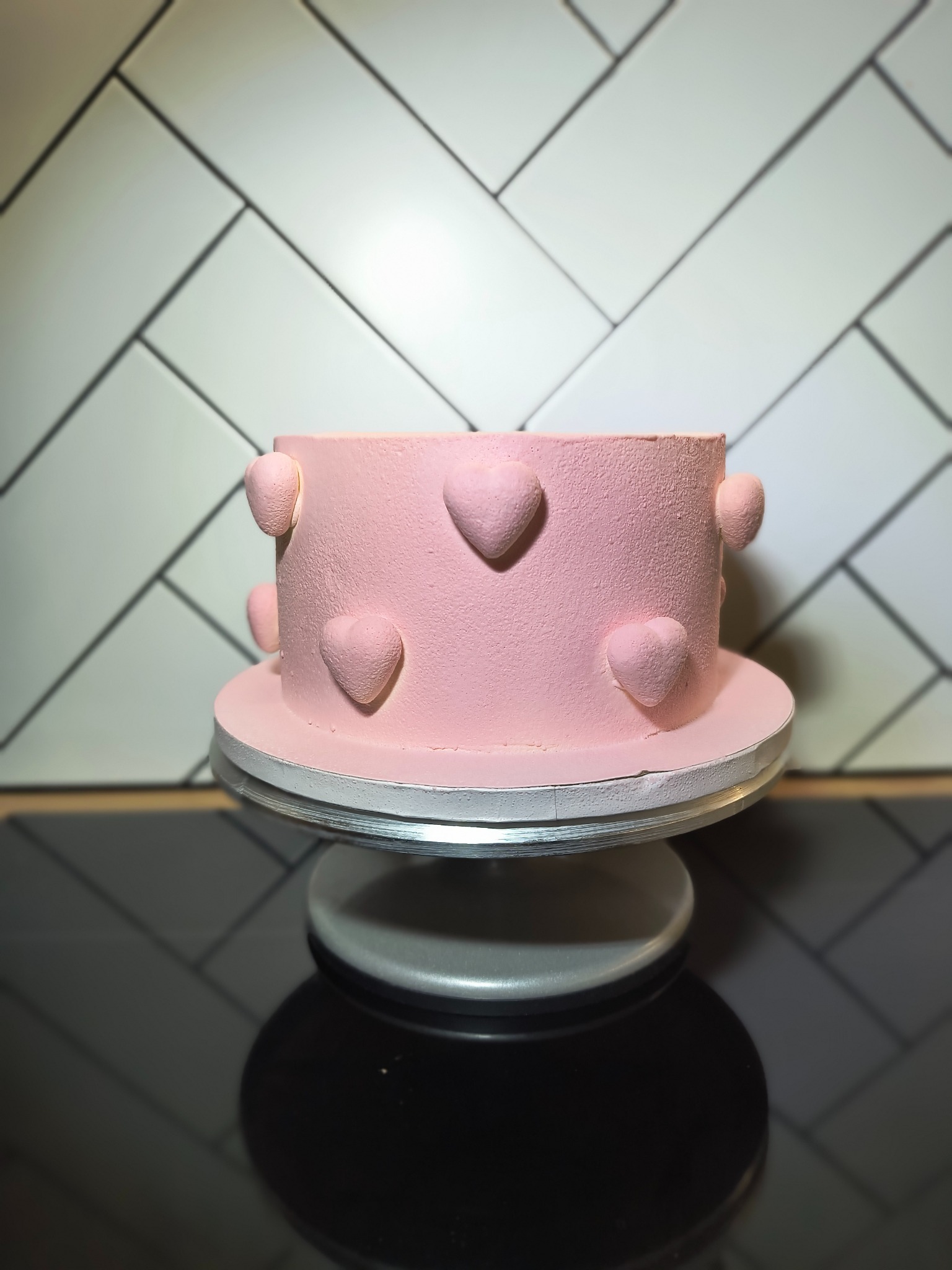 Różowy tort z serduszkami na obrotowej podstawie, ustawiony na czarnym blacie, z tłem z białych płytek ułożonych w jodełkę.