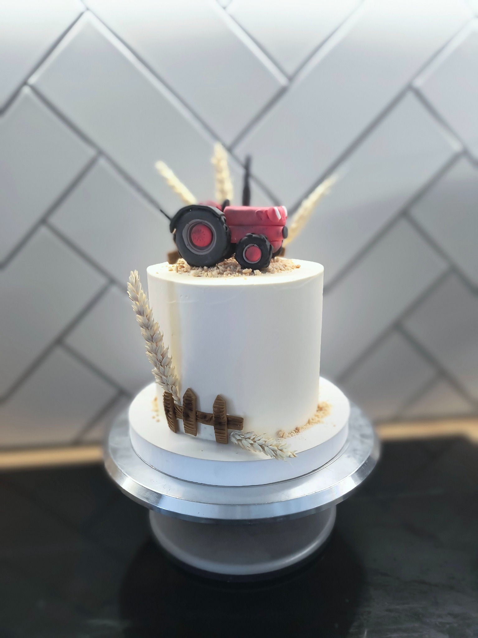 Tort z motywem traktora: biały krem, czerwony traktor z masy cukrowej, dekoracje z pszenicy i płotka. Ustawiony na srebrnej paterze, tło z białych płytek.