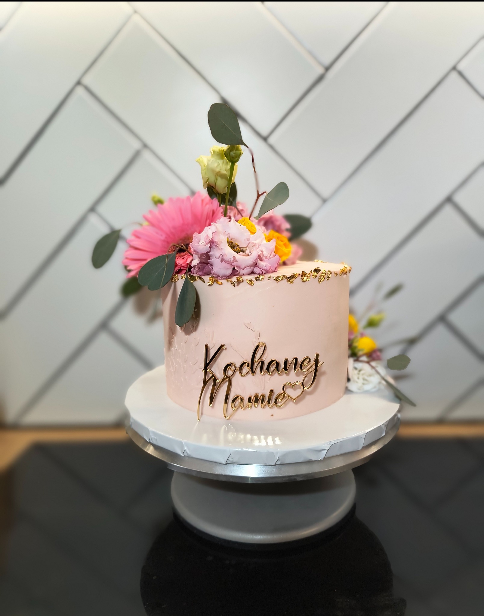 Różowy tort 'Kochanej Mamie' z kwiatami i złotymi płatkami, na paterze, tło z białych płytek. Dekoracja okolicznościowa.