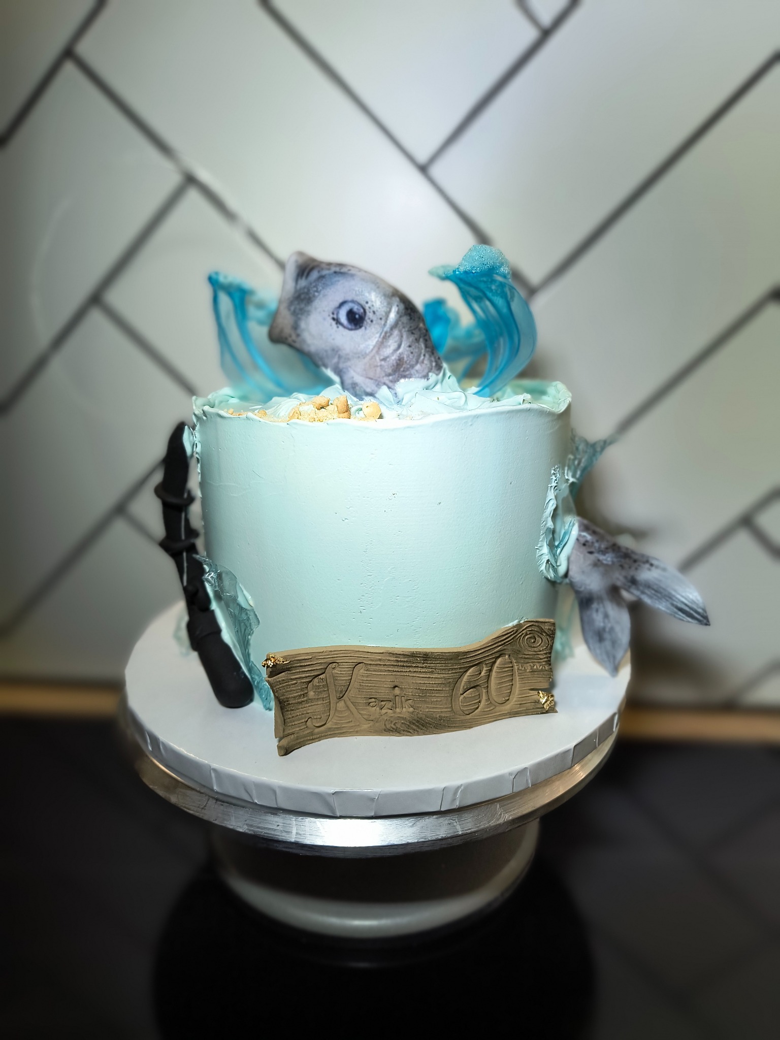 Niebieski tort urodzinowy z motywem ryby i napisem 'Kaziu 60', na obrotowej podstawie, z tłem w biało-szarą kratkę. Dekoracja cukiernicza.