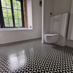 Wykończona łazienka z geometryczną czarno-białą podłogą, wiszącą toaletą i oknem z widokiem na zieleń.