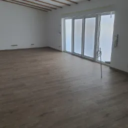 Przestronne, jasne wnętrze z drewnianą podłogą i sufitowymi belkami, duże okna częściowo zasłonięte folią, widoczne punkty instalacji elektrycznej w ścianach.