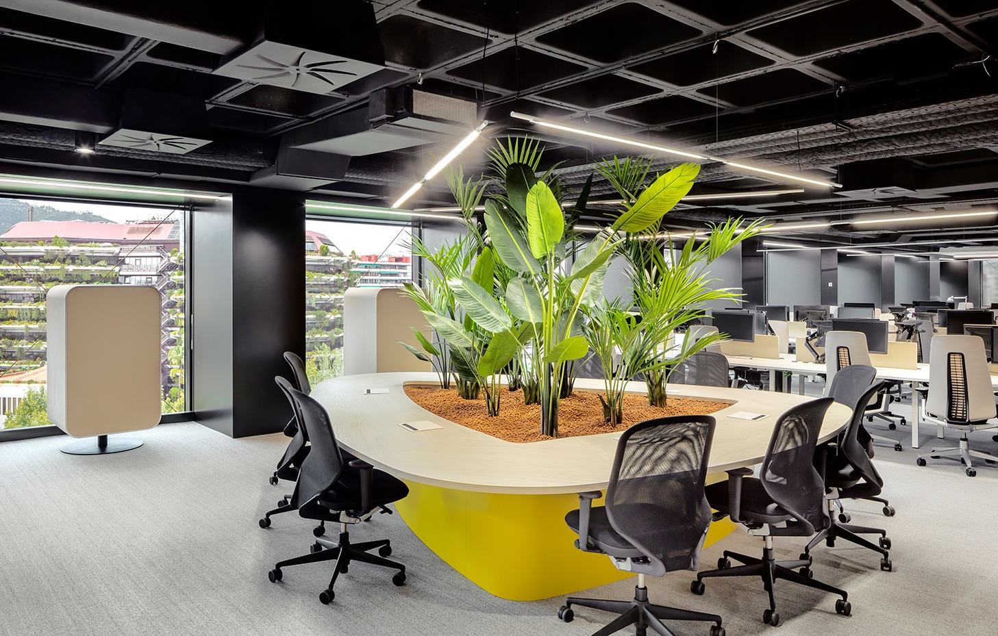 Nowoczesne biuro z systemem wentylacji sufitowej, stołem konferencyjnym z roślinami i widokiem na zielony budynek przez okno.