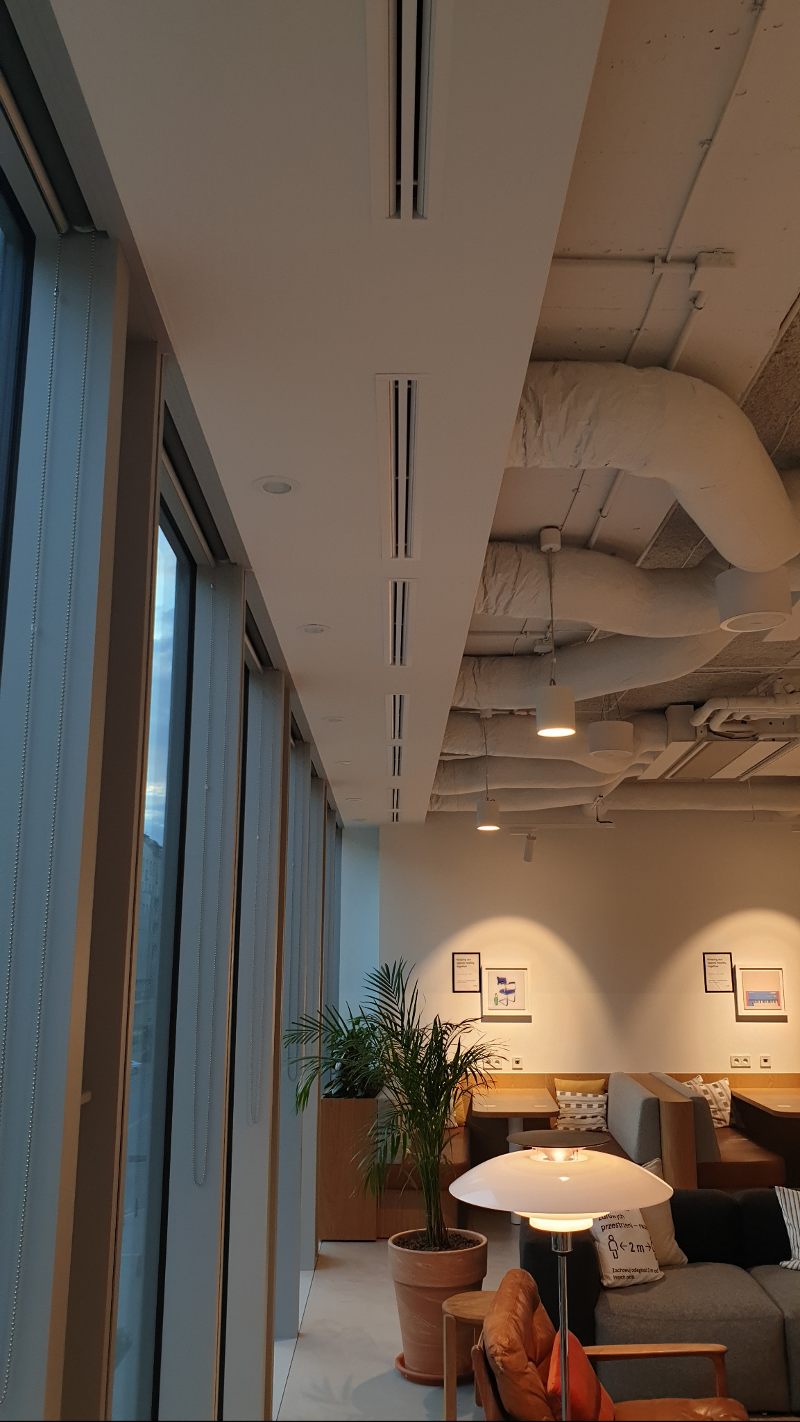 Wnętrze biura z widocznymi liniowymi nawiewnikami wentylacyjnymi w suficie, dużymi oknami z żaluzjami i widocznymi instalacjami pod sufitem.