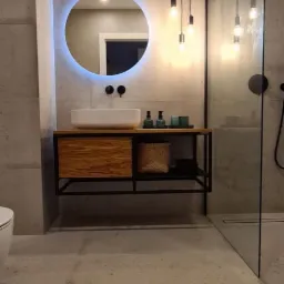 Nowoczesna łazienka z betonowymi płytkami, okrągłym lustrem z podświetleniem LED, drewnianą szafką pod umywalką i szklaną kabiną prysznicową z czarną armaturą oraz wiszącymi lampami z żarówkami...