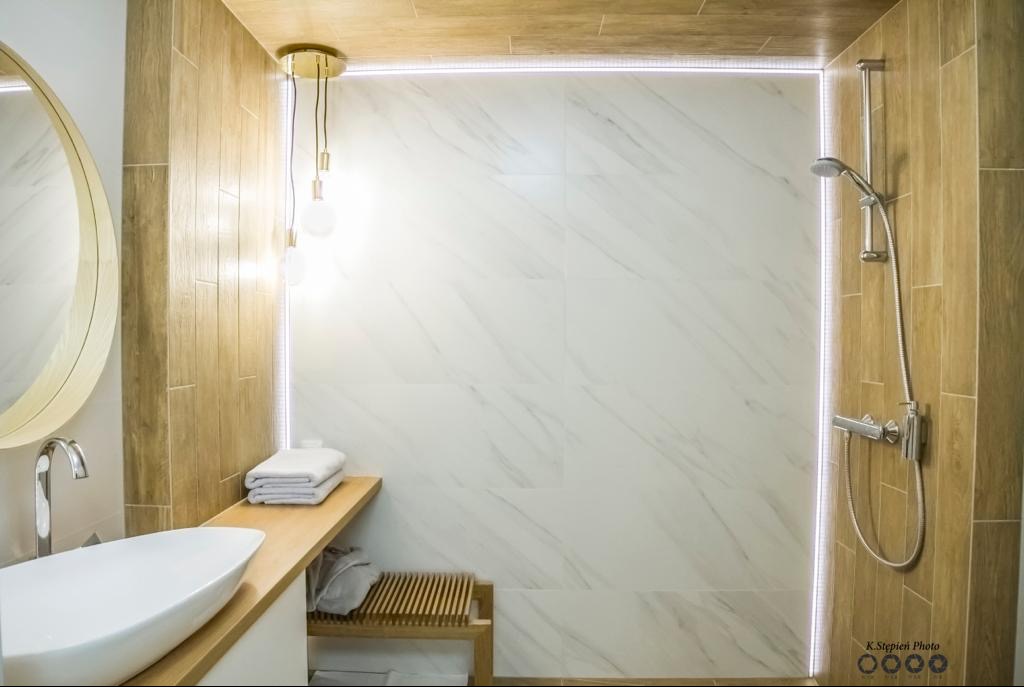 Nowoczesna łazienka z białymi marmurowymi płytkami, drewnopodobnymi panelami ściennymi, okrągłym lustrem z podświetleniem LED, wiszącą lampą z trzema żarówkami i chromowaną armaturą prysznicową.