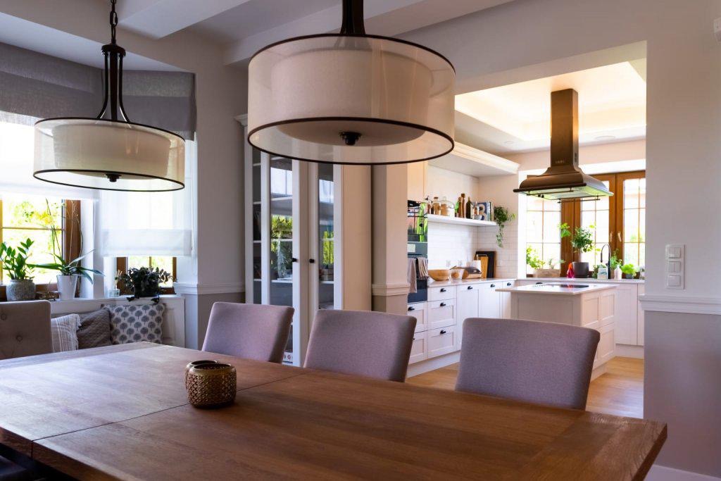 Jasne wnętrze jadalni połączonej z kuchnią, drewniany stół z szarymi krzesłami, wiszące lampy z abażurami, zabudowa kuchenna w bieli z drewnianymi oknami w tle.