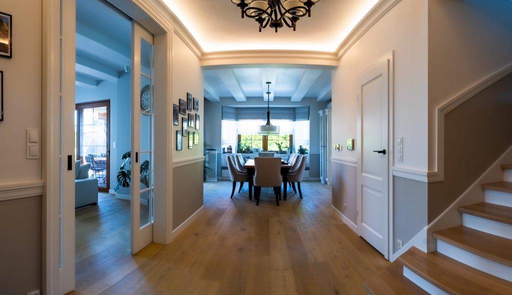 Przestronny korytarz z drewnianą podłogą prowadzący do jadalni, białe ściany z szarym pasem, drzwi przesuwne po lewej, schody po prawej, dekoracyjne oświetlenie sufitowe.