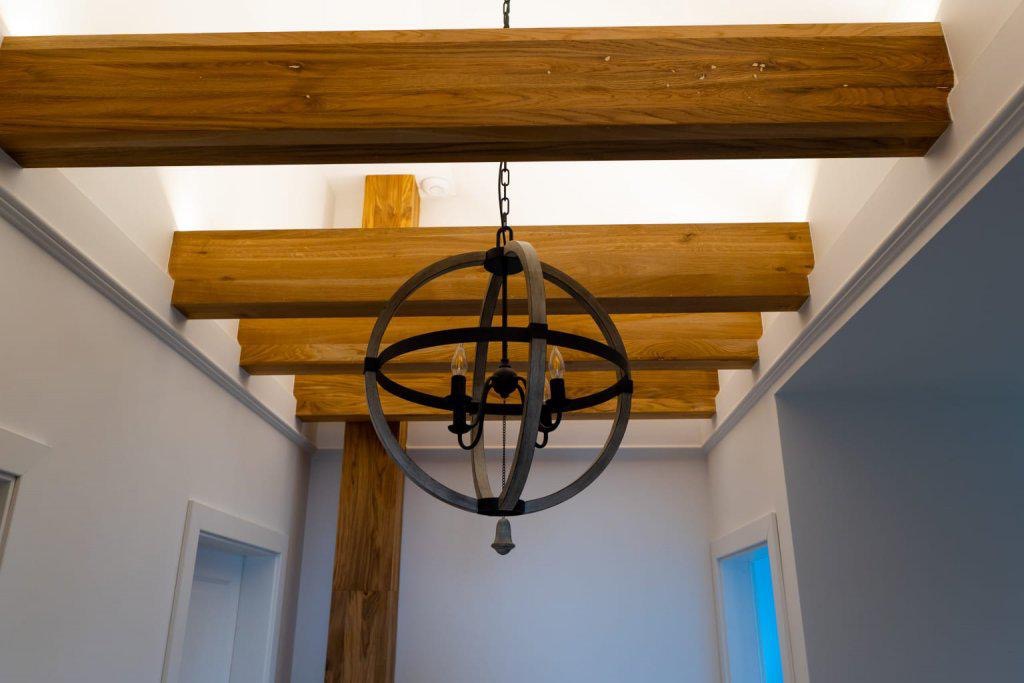 Drewniane belki stropowe z oświetleniem LED, uzupełnione dekoracyjnym żyrandolem w kształcie kuli, w jasnym wnętrzu.
