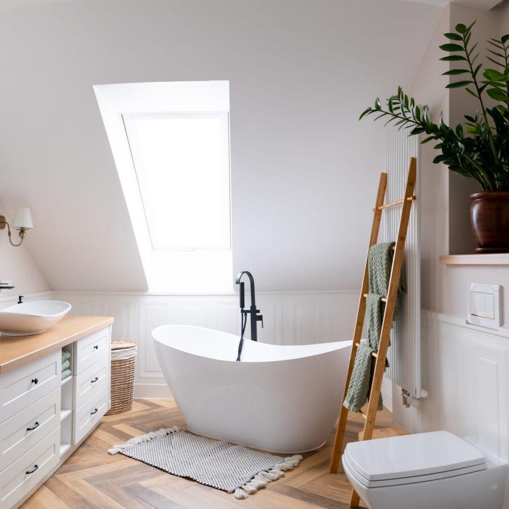 Jasna łazienka na poddaszu z wolnostojącą wanną, drewnianą drabinką z ręcznikami, umywalką nablatową i oknem dachowym.