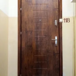 Brązowe drzwi wejściowe z okleiną imitującą drewno, wyposażone w wizjer i srebrny zamek, zamontowane w korytarzu bloku mieszkalnego, obok numeru mieszkania 118.