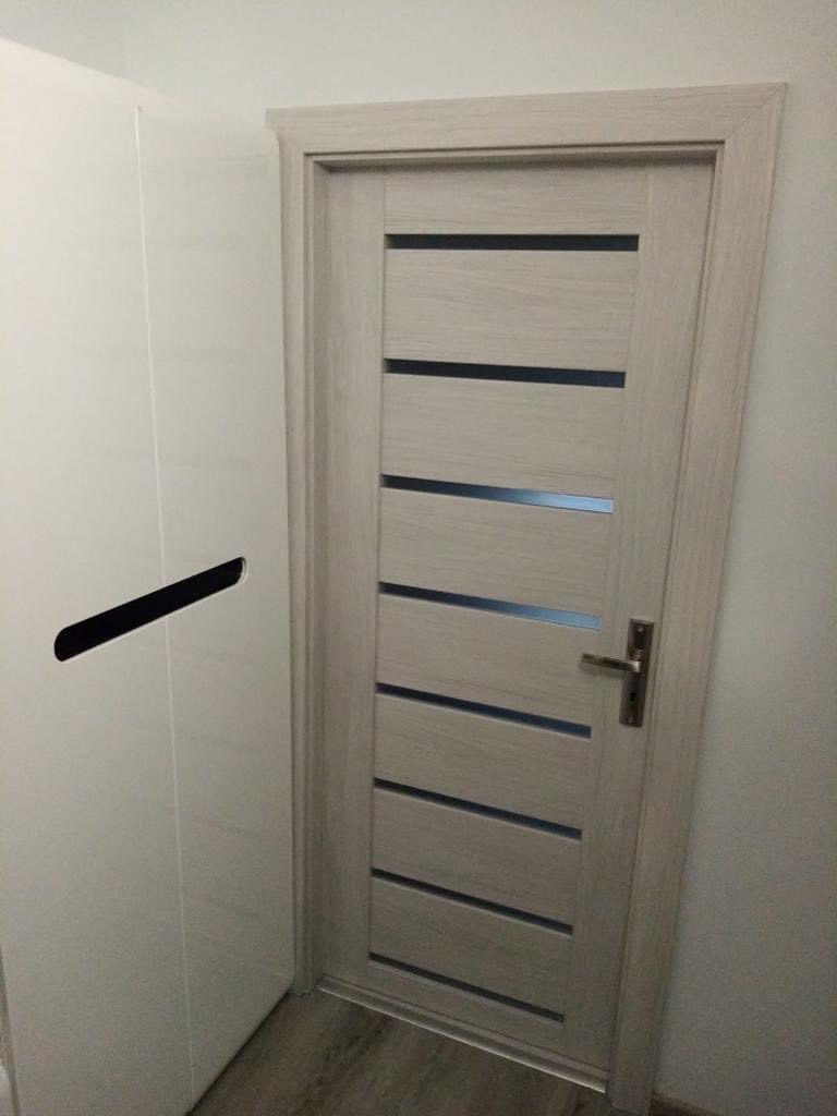 Jasnoszare skrzydło drzwiowe z poziomymi przeszkleniami, widoczna klamka i fragment szafy w tle.