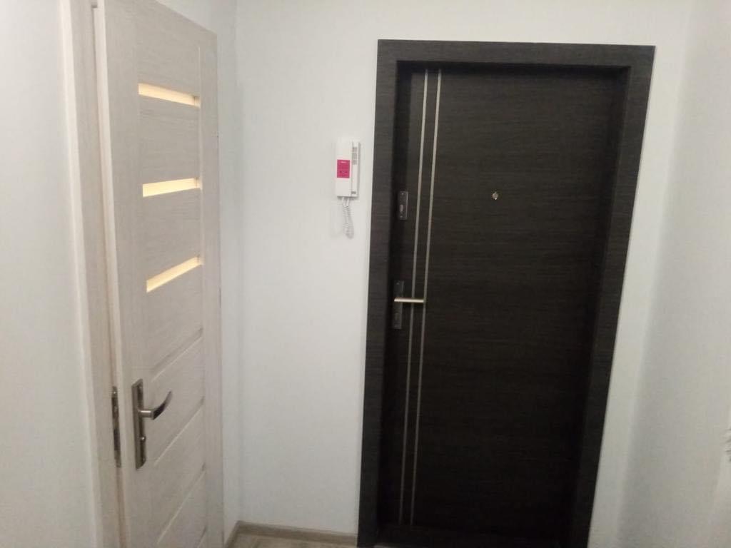 Dwa rodzaje drzwi w korytarzu: jasne, z poziomymi przeszkleniami i ciemne, z metalowymi paskami, zamontowane obok domofonu.