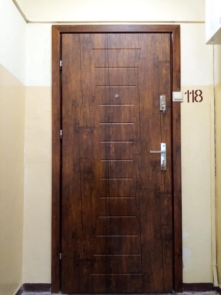 Brązowe drzwi wejściowe z okleiną imitującą drewno, wyposażone w wizjer i srebrny zamek, zamontowane w korytarzu bloku mieszkalnego, obok numeru mieszkania 118.