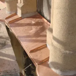 Detal montażu miedzianej obróbki blacharskiej na elewacji budynku, widoczne rusztowanie i elementy wykończeniowe z bliska.