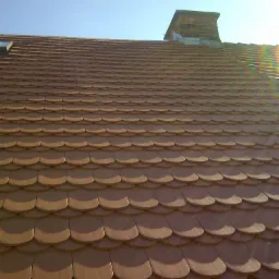 Dach pokryty brązową dachówką karpiówką, widoczny komin z cegły oraz okno dachowe, całość oświetlona mocnym słońcem z efektem tęczy.