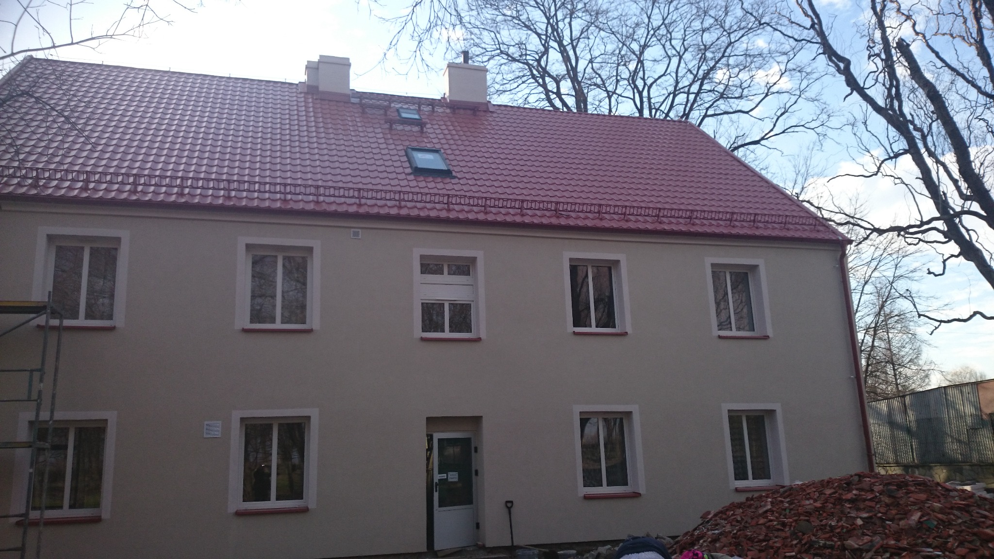 Budynek dwukondygnacyjny z nowym, czerwonym dachem z dachówką, ocieploną elewacją w kolorze beżowym, widoczne okna z białymi ramami, częściowo rozstawione rusztowanie z lewej strony, oraz sterta...