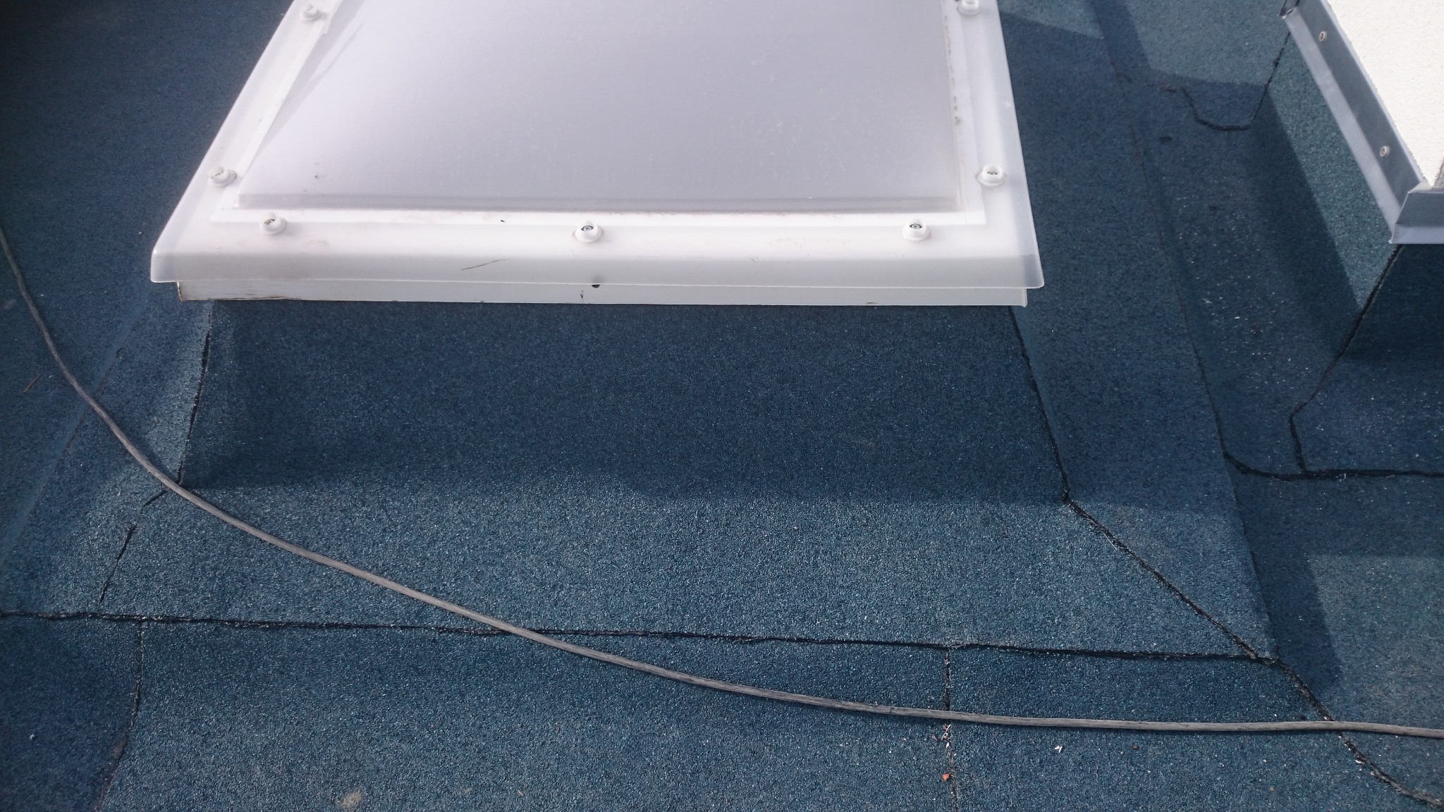 Widok z góry na fragment dachu pokrytego papą w kolorze granatowym, z zamontowanym świetlikiem dachowym o białej ramie i widocznym przewodem elektrycznym biegnącym po powierzchni dachu.