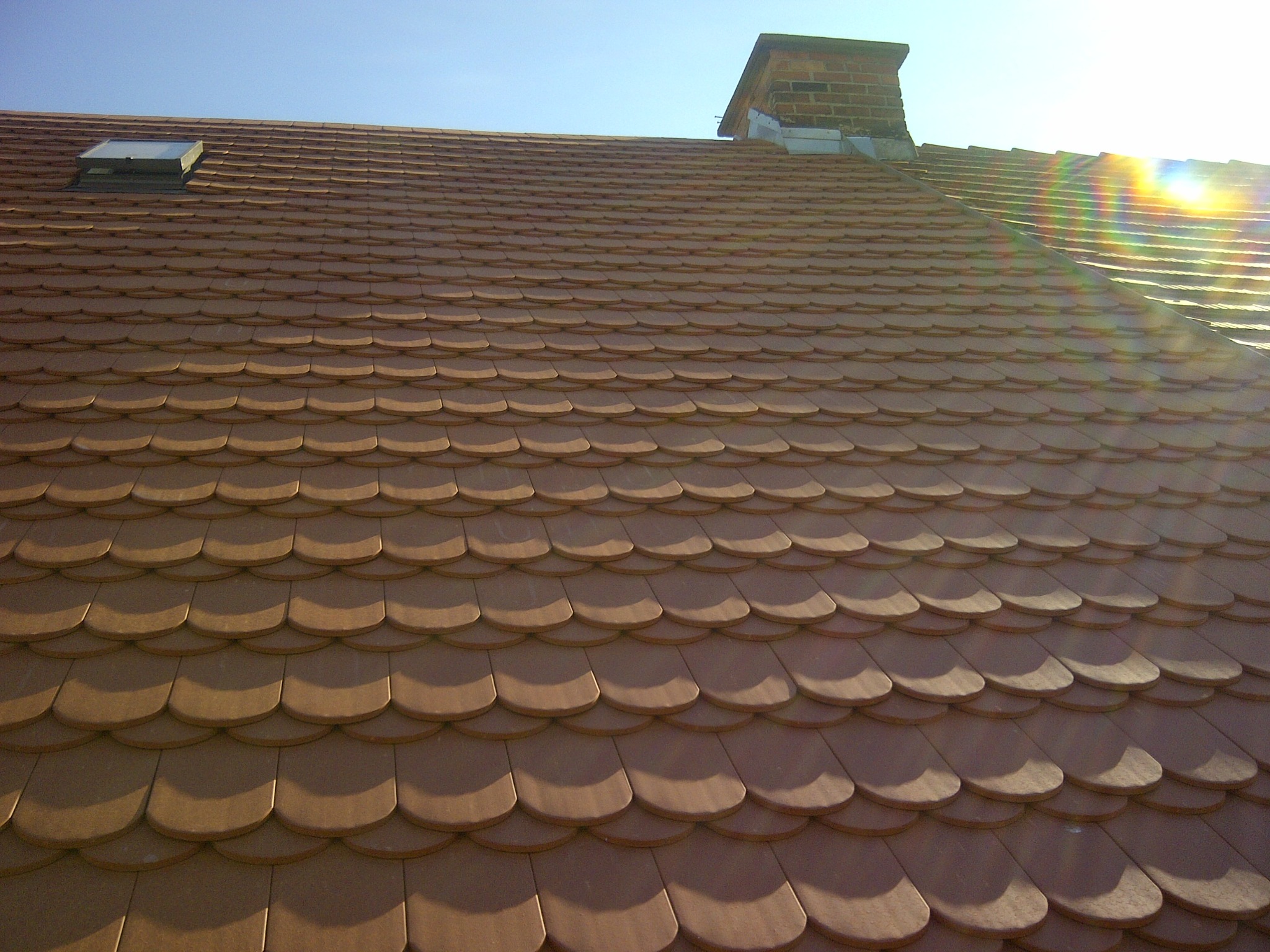 Dach pokryty brązową dachówką karpiówką, widoczny komin z cegły oraz okno dachowe, całość oświetlona mocnym słońcem z efektem tęczy.