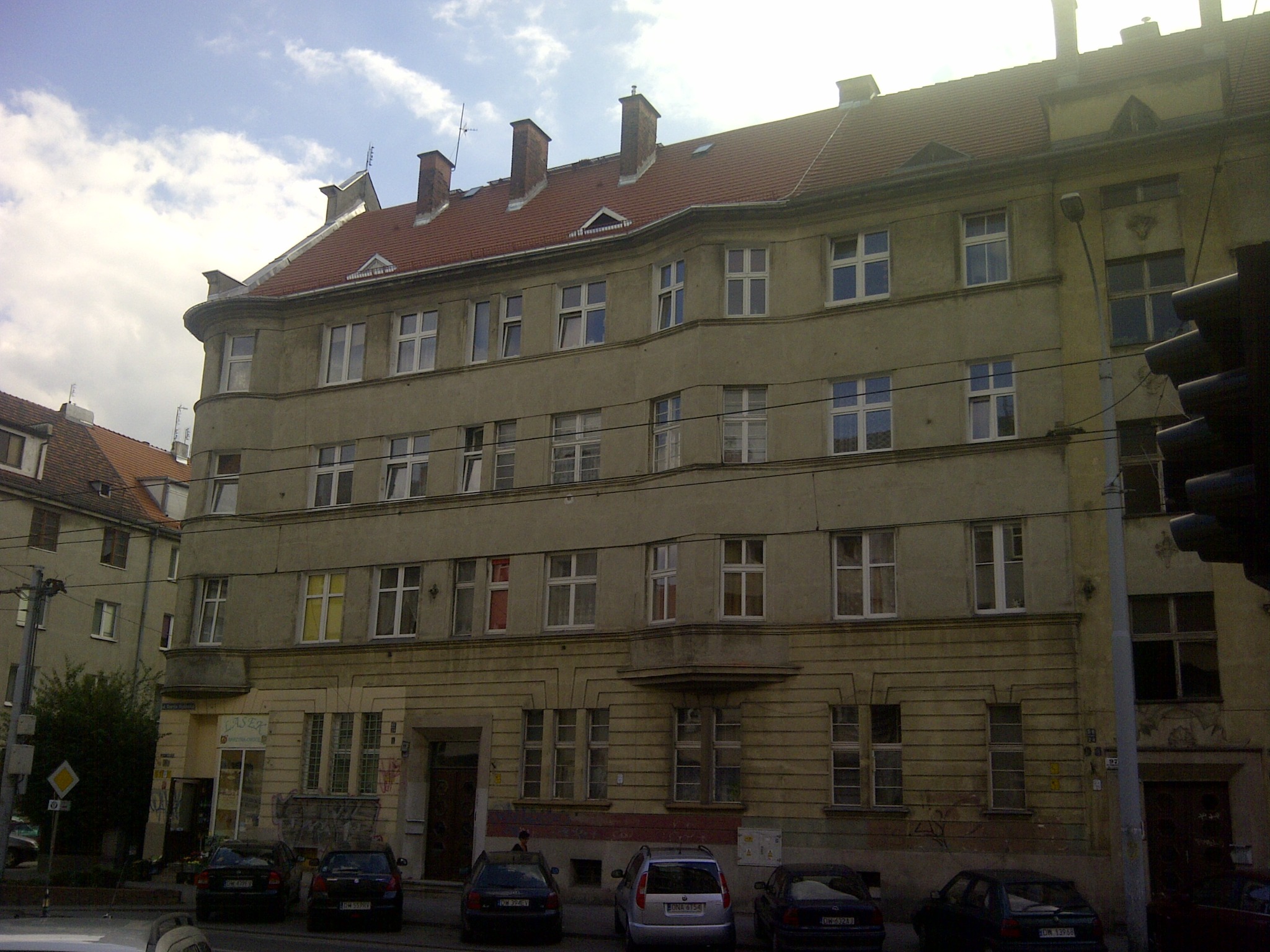 Wielopiętrowy budynek mieszkalny z czerwonym dachem, widoczny z poziomu ulicy, z zaparkowanymi samochodami przed budynkiem i sygnalizacją świetlną po prawej stronie.