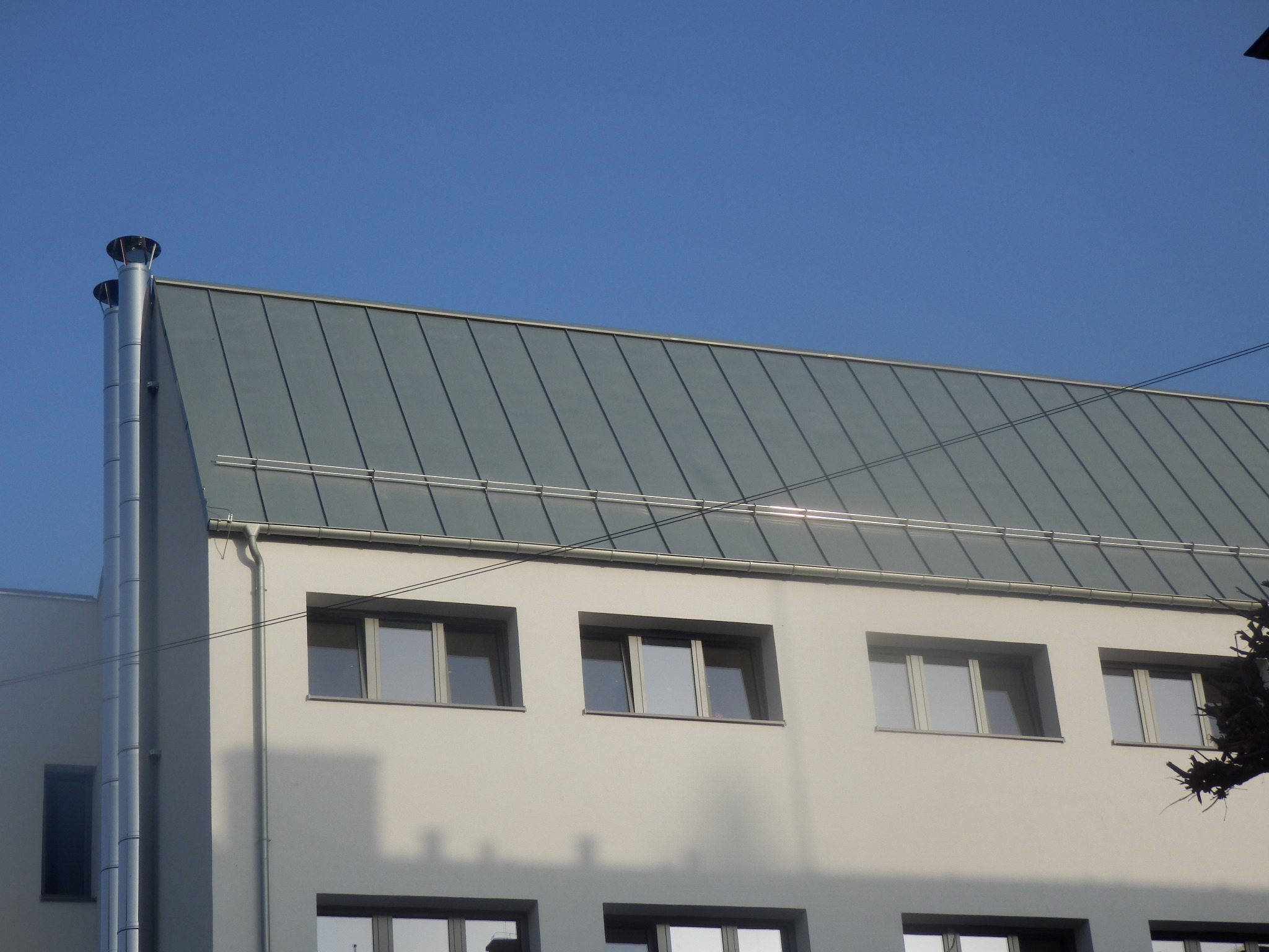 Szary dach z blachy na budynku z jasną elewacją, widoczne okna i kominy wentylacyjne.