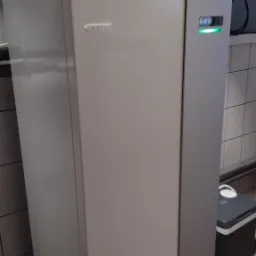 Biała jednostka wewnętrzna z widocznym logo Nibe i zieloną diodą, z instalacją hydrauliczną na górze, stojąca w pomieszczeniu wyłożonym kafelkami.