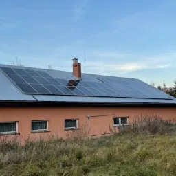 Instalacja fotowoltaiczna na dachu budynku z blachy falistej, widok z dołu na tle pochmurnego nieba i drzew iglastych.