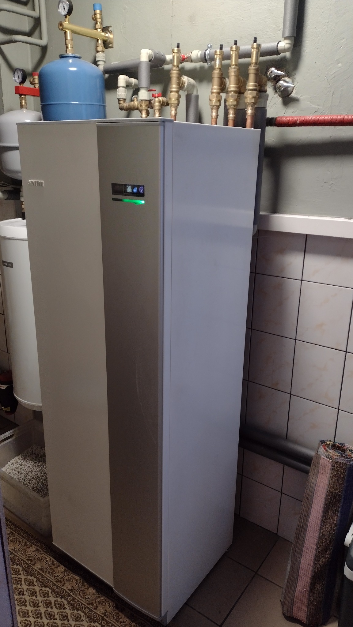 Szara jednostka wewnętrzna z zielonym wskaźnikiem LED, podłączona do instalacji hydraulicznej w kotłowni wykończonej kafelkami.